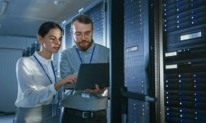 Deux personnes travaillent dans un data center, examinant des informations sur un ordinateur portable devant une rangée de serveurs.