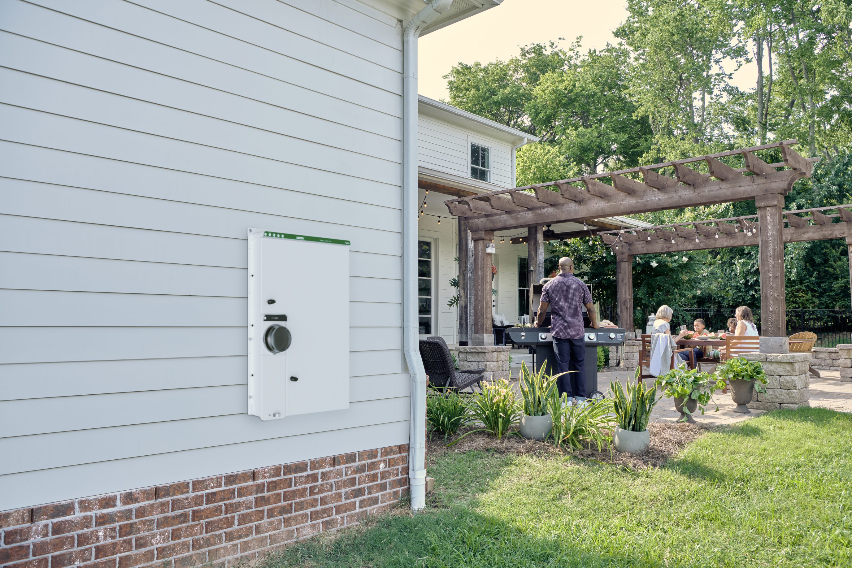 image of a wiser energy center product fixed on to the sife of a suburban house. in the background there is a family having a barbeque.