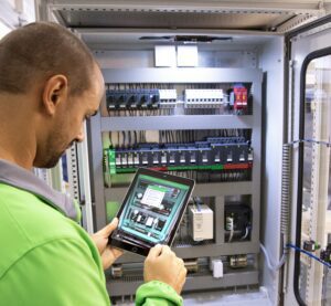 an engineer holding a tablet which monitors machine perfoemance. he is standing infront of a tesys island control panel