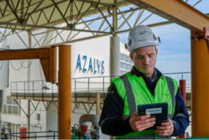 A Schneider Electric worker wearing a green safety vest stands in front of the Azalys Suez building holding a tablet.