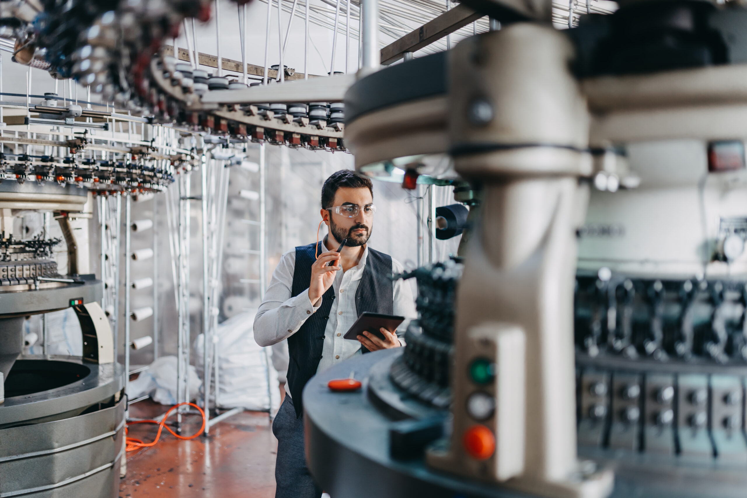 A man, centered in the frame, and holding a tablet is surrounded by machinery in a factory.