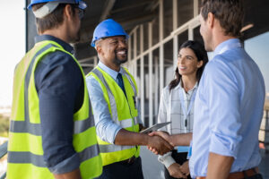 4 people, 2 engineers and 2 business people, stand together in front of a building. An engineer and businessman shake hands.