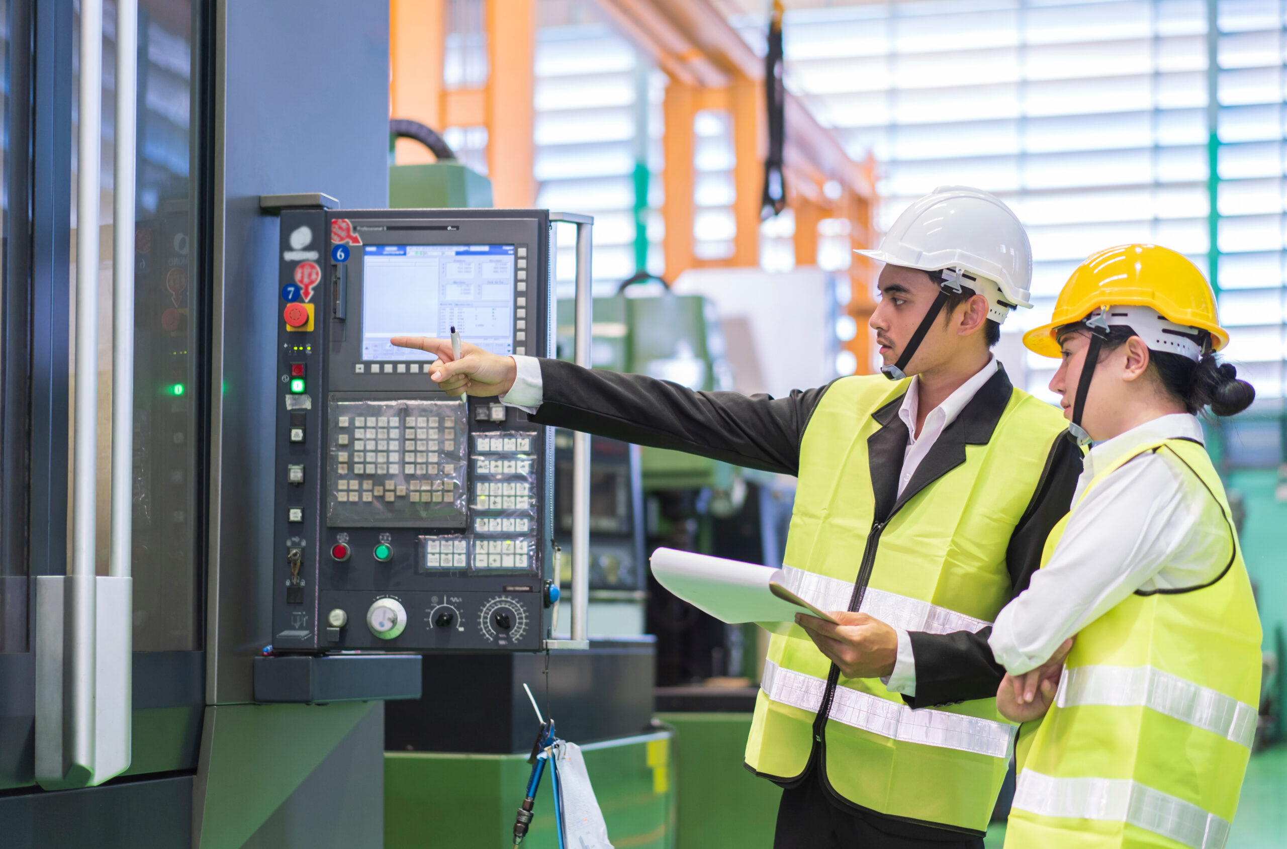 A man and woman wearing yellow safety vests and hard hats look at a power panel in an industrial setting