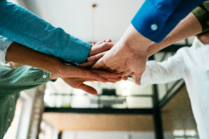 The hands of 6 different people are put together during a huddle in an office workspace setting.