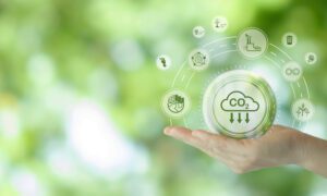 A hand is extended, palm-up, against a blurred green background, with a graphic overlay illustrating various environmental icons encircling a central 'CO2' cloud symbol, representing the concept of 'Scope 3 emissions' in greenhouse gas accounting, which include indirect emissions in a company's value chain.