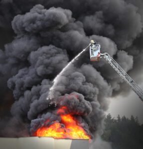 A firefighter atop an extended ladder from a fire truck douses a massive fire with water. Thick black smoke billows from the blazing inferno against a backdrop of a grey sky, highlighting the importance of fire alarm safety in preventing such hazardous situations.