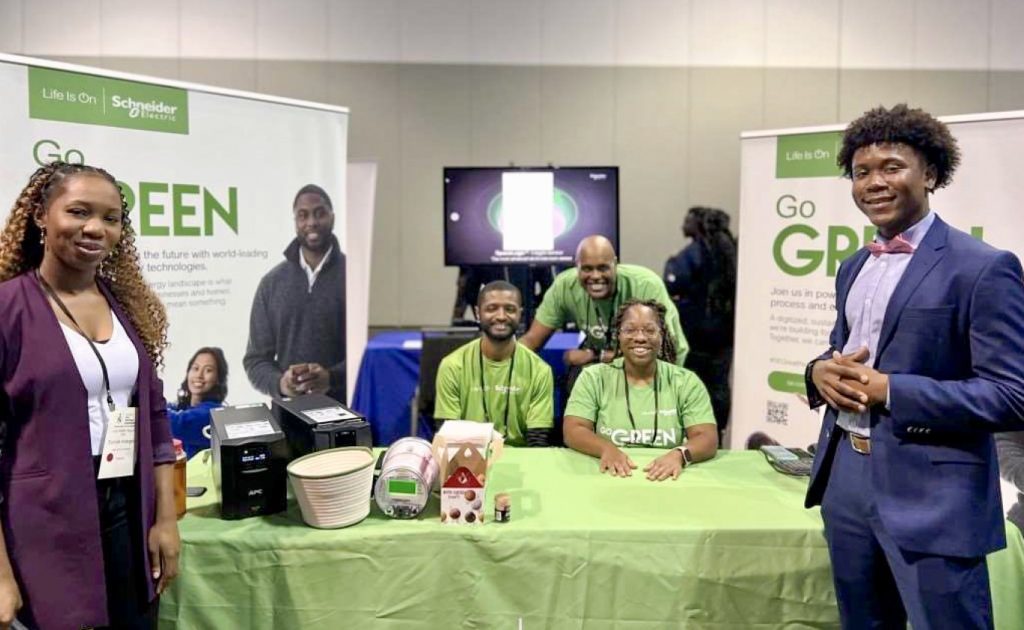 This image shows three Schneider Electric employees at a table smiling.  There are two NSBE students standing next to the table.