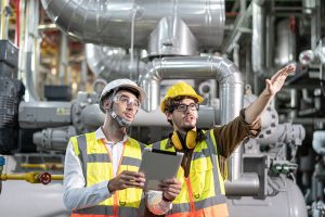 Two workers in safety vests and hardhats discuss equipment in a manufacturing plant.