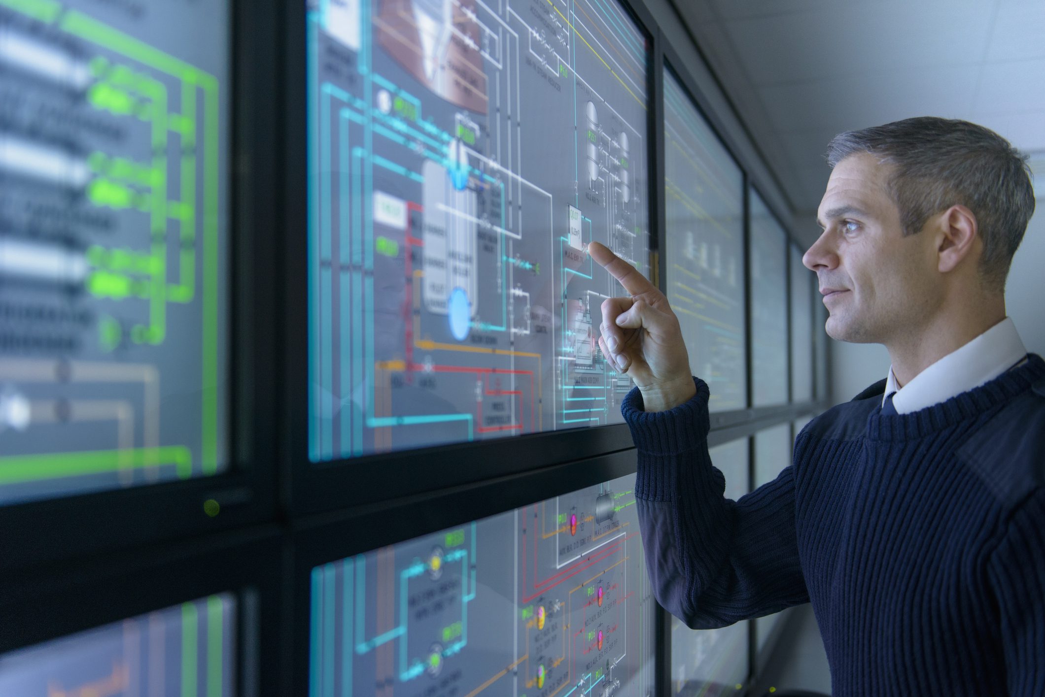 Person using touch screen in operations control room, part of a network of screens with graphical interfaces.