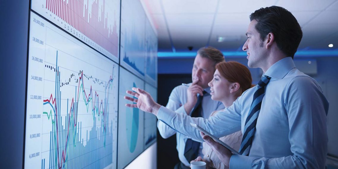 A group of business people looking at charts on a wall of computer screens.