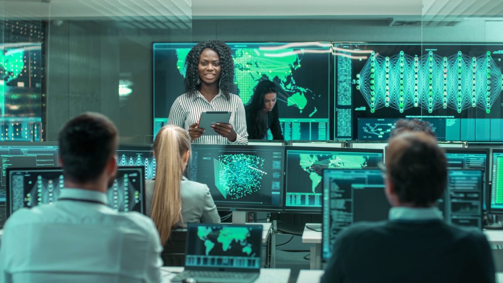 A chief project engineer consults a tablet computer and holds a briefing for a team of scientists that are working at coordinated display screens.