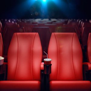 An image of a movie theater with plush red seats in the foreground, focused on a singular seat with a popcorn box and a soda cup in the cup holder. The darkened theater setting suggests a private viewing, with the screen barely visible in the background, bathed in a soft, glowing light. The scene is set for the screening of 'The End We Start From', a film that underscores the pressing need for climate action through its narrative, urging viewers to consider the impact of environmental change on personal and collective futures.