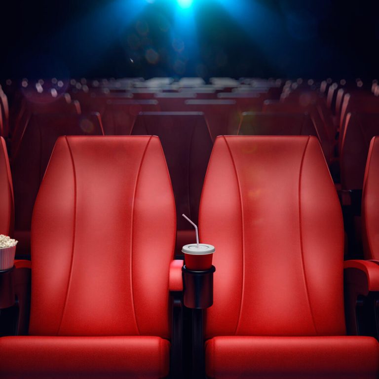 An image of a movie theater with plush red seats in the foreground, focused on a singular seat with a popcorn box and a soda cup in the cup holder. The darkened theater setting suggests a private viewing, with the screen barely visible in the background, bathed in a soft, glowing light. The scene is set for the screening of 'The End We Start From', a film that underscores the pressing need for climate action through its narrative, urging viewers to consider the impact of environmental change on personal and collective futures.