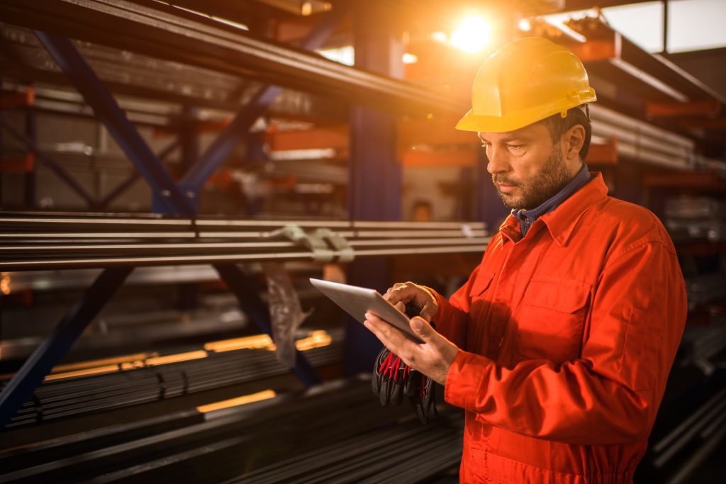 Adult male worker wearing red coveralls standing in aluminum mill and reading on a tablet. 