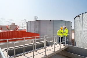 Two workers in yellow safety jackets stand on a catwalk between water reservoirs