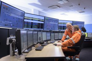 Two workers in orange suits monitor multiple screens with data and control panels in a modern control room.