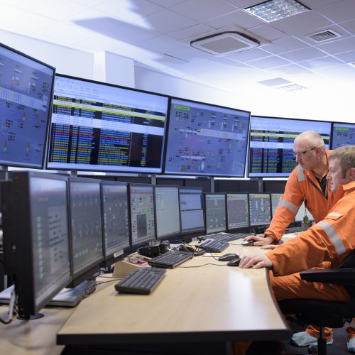 Two workers in orange suits monitor multiple screens with data and control panels in a modern control room.