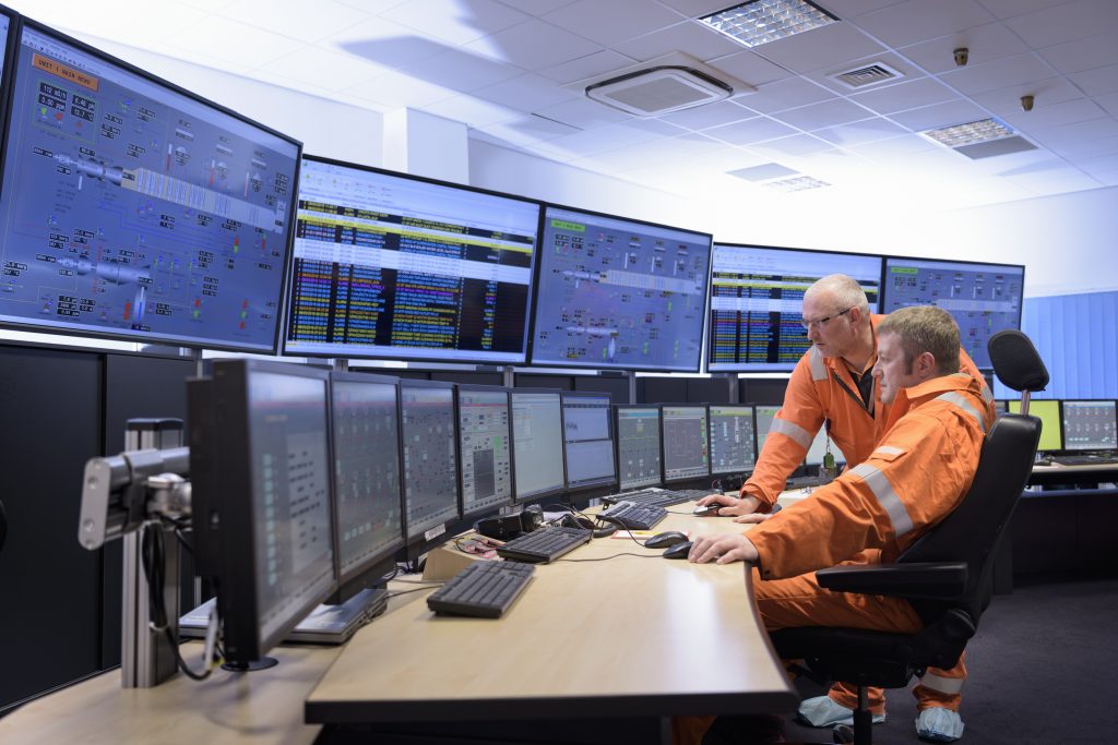 Two workers in orange suits monitor multiple screens with data and control panels in a modern control room.