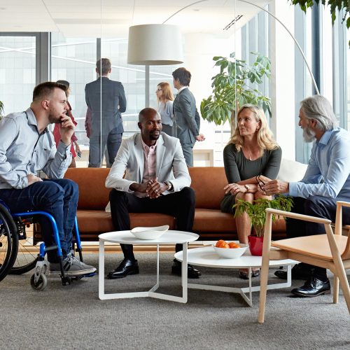 A group of people sitting in chairs indoors, surrounded by furniture including a coffee table and houseplant. The setting has windows that let in natural light. Among the individuals present, there is one person using a wheelchair. The environment appears cozy, with a focus on the interactions among the group.