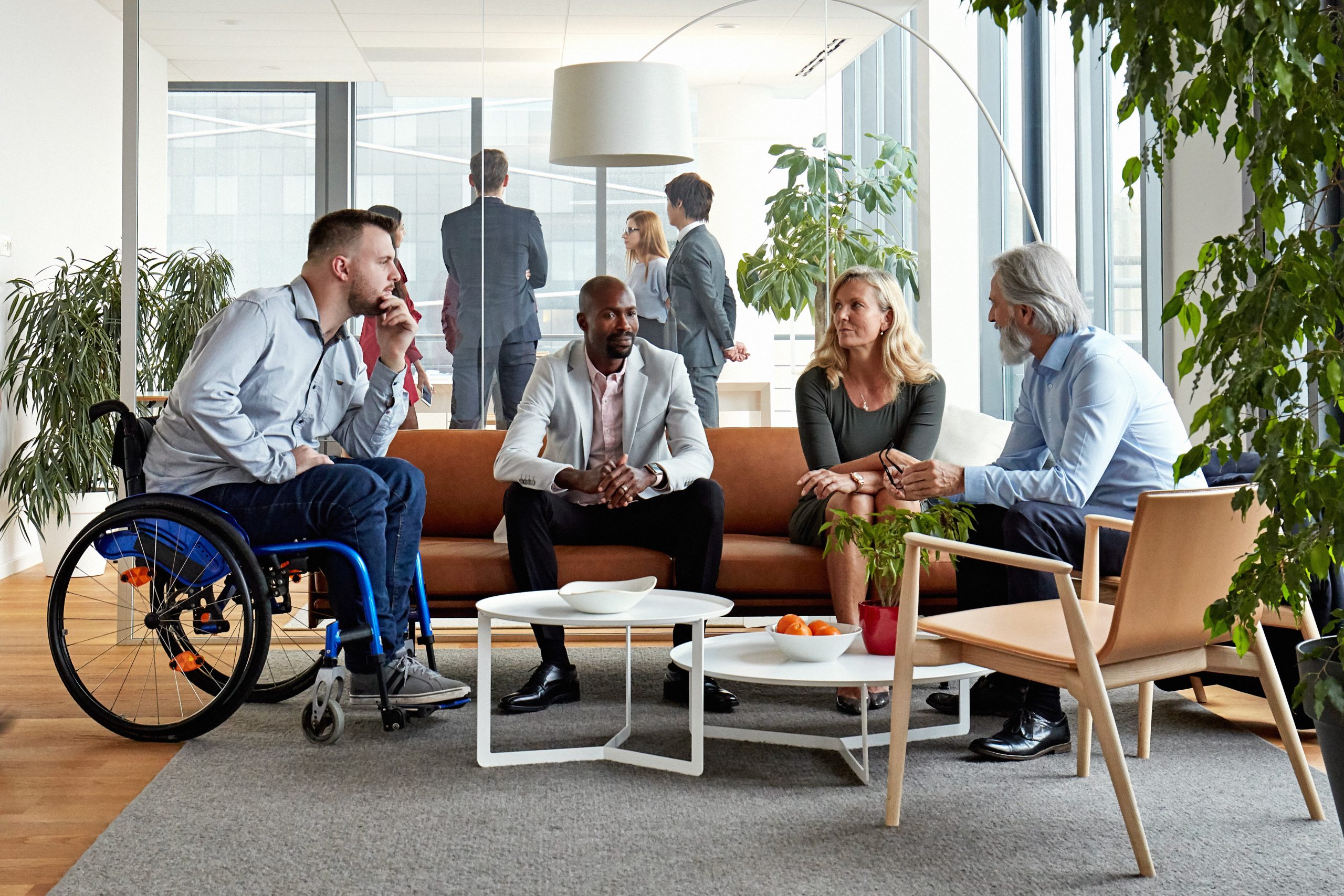 A group of people sitting in chairs indoors, surrounded by furniture including a coffee table and houseplant. The setting has windows that let in natural light. Among the individuals present, there is one person using a wheelchair. The environment appears cozy, with a focus on the interactions among the group.