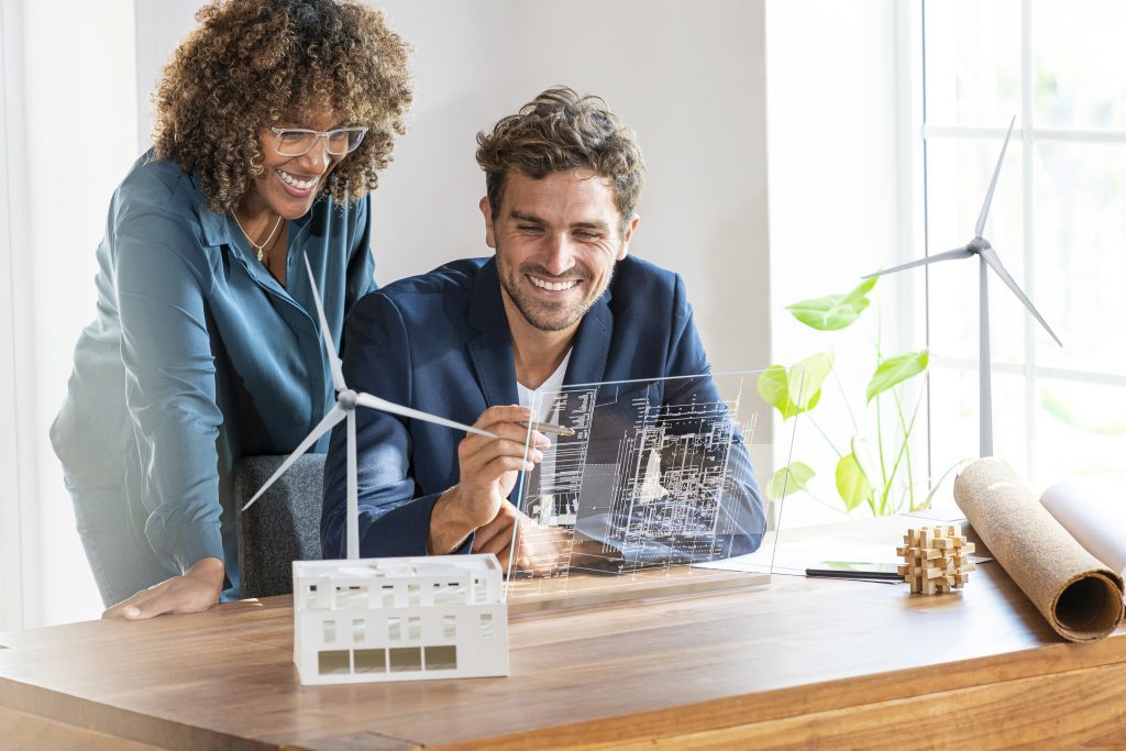 Alt-text: Two professionals, a woman with curly hair and glasses and a man in a blue suit, smile while examining a transparent display showing building schematics. On the desk are models of a wind turbine and a building, along with other project materials. The scene suggests they are Smart Grid EcoXperts collaborating on an eco-friendly architectural or energy project.