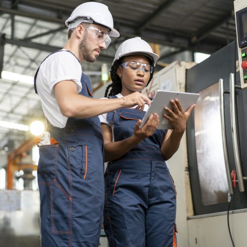 Two workers in overalls look at a data on a tablet in a factory.