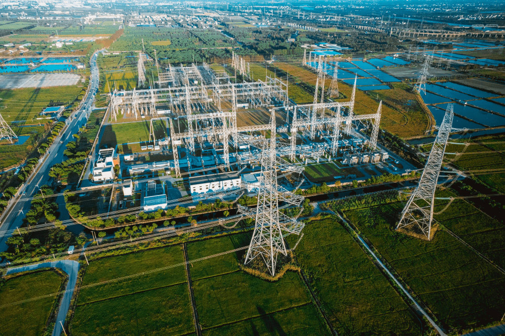 Aerial view of a large electrical substation in a rural area. The substation is filled with transformers, power lines, and electrical equipment. Surrounding the substation are green fields, roads, and in the distance, a mix of urban and industrial development. Several large transmission towers are visible, carrying high-voltage power lines away from the substation.