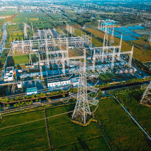 Aerial view of a large electrical substation in a rural area. The substation is filled with transformers, power lines, and electrical equipment. Surrounding the substation are green fields, roads, and in the distance, a mix of urban and industrial development. Several large transmission towers are visible, carrying high-voltage power lines away from the substation.