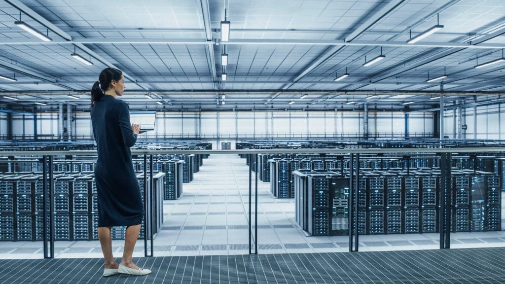 Data Center Engineer Using Laptop Computer standing in front a row of AI-ready data centers. Server Farm Cloud Computing Specialist Facility with Multiethnic Female System Administrator Working with Data Protection Network for Cyber Security.