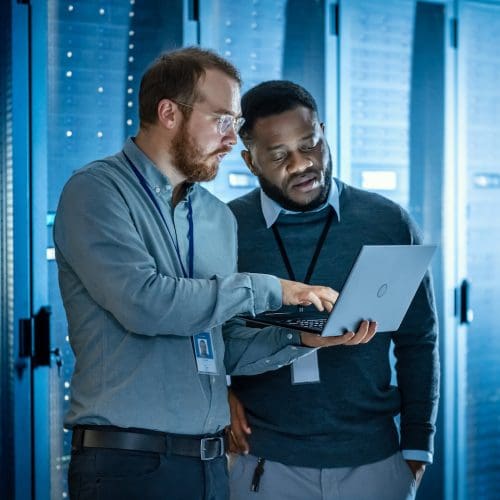 Bearded IT Technician in Glasses with Laptop Computer and Black Male Engineer Colleague are Using Laptop in Data Center while Working Next to Server Racks. Running Diagnostics or Doing Maintenance Work